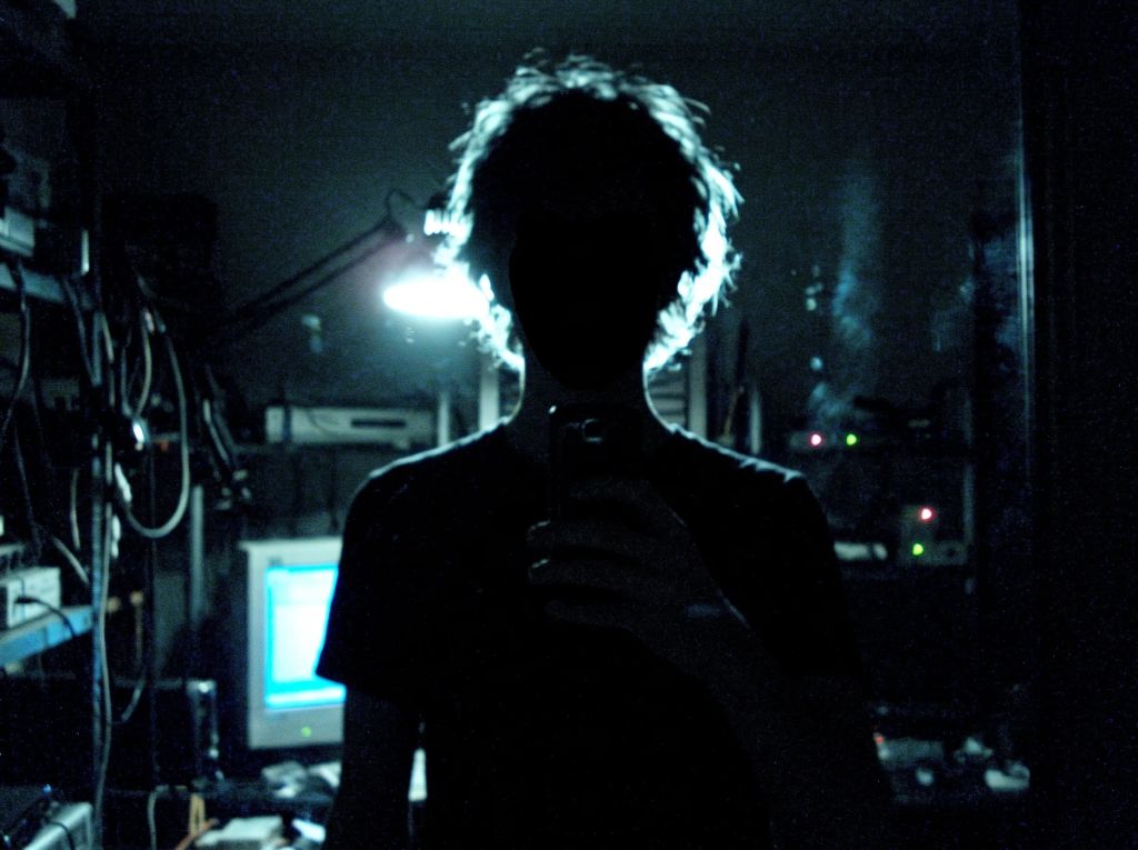 backlit mirror selfie, face completely in shadow, wild hair haloed by a lamp, old equipment in the dark behind